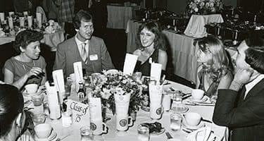 An old black and white photo of alumni sitting and eating around a table